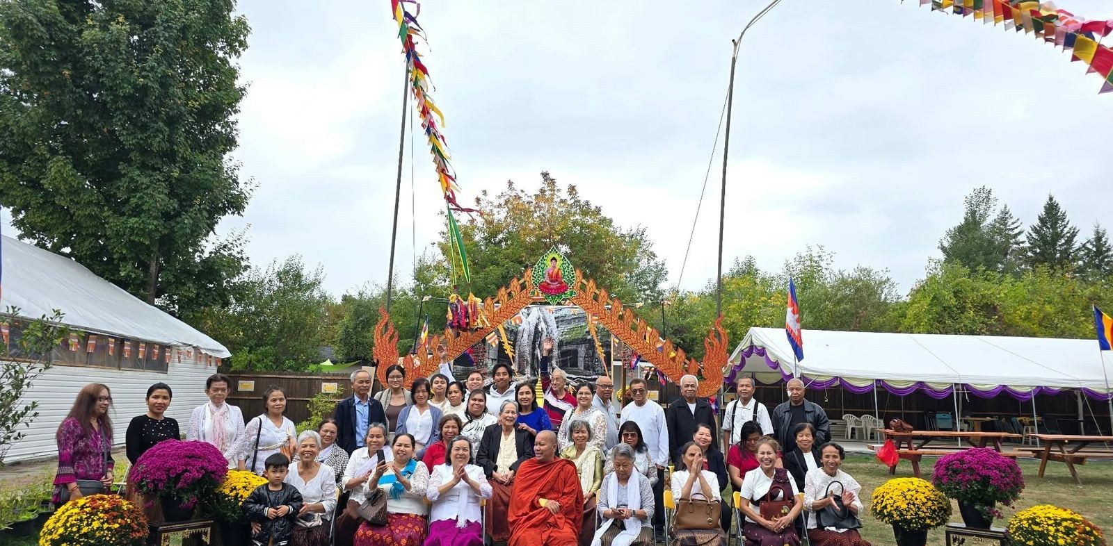 A monk and community members seated in front of a decorated gateway, with white tents and pots of chrysanthemums